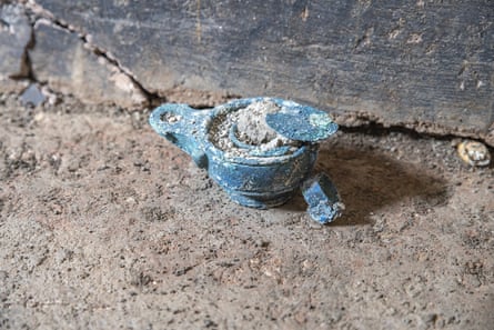 An oil lamp with a spout, corroded blue, sits on the stone floor of a dig site