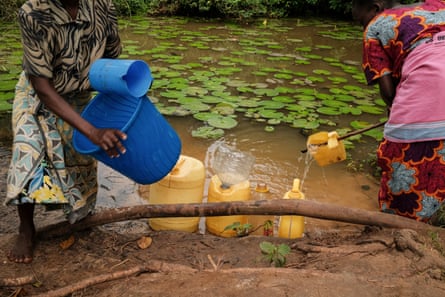 Two women stand on a muddy bank and fill brightly coloured buckets and containers
