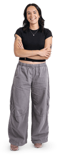 A dark-haired woman stands, arms crossed, wearing baggy grey trousers and a black crop top