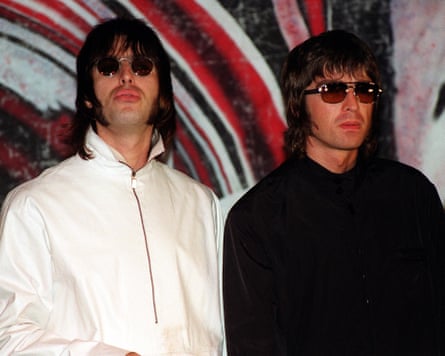 Liam and Noel Gallagher pose for a photo in 1999. They both wear sunglasses and have collar-length hair with long fringes. Liam wears a white zip-up anorak and Noel a black jacket. They stand in front of a swirling red, white and black backdrop