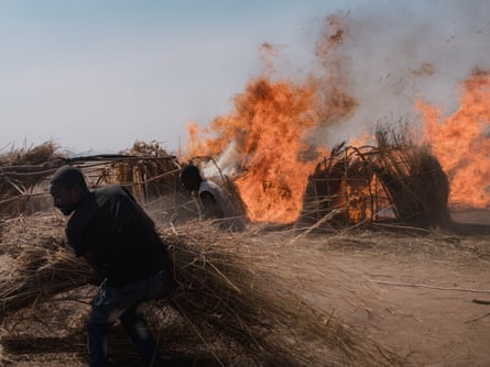 A man shelters while a fire burns palm huts on dusty land.