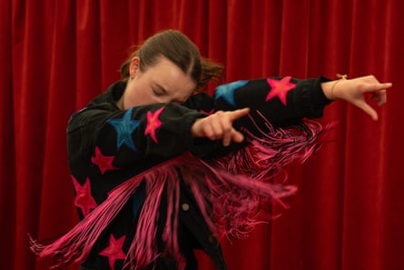 Bella Ramsey mid-dance move, with both arms stretched out to her left, her index figures pointing, wearing a black outfit with red and blue star on and red tassels on the arms