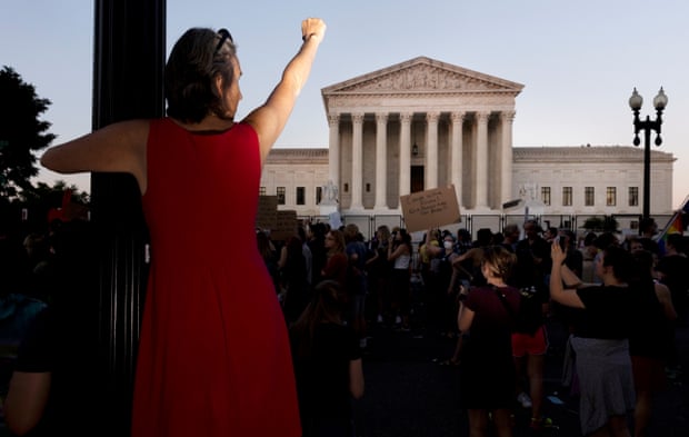 A woman in a red dress clings to a pole with one fist in the air aimed at the supreme court building in Washington DC.