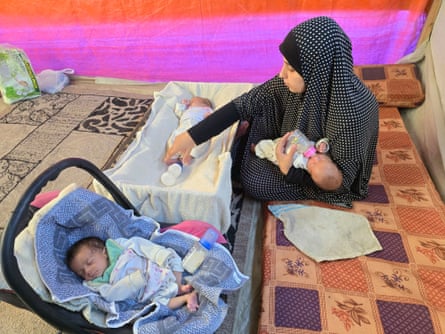 Woman in a hijab trying to feed three tiny babies with bottles – one in her arms, one in a crib, one in a carrier – in a tent.