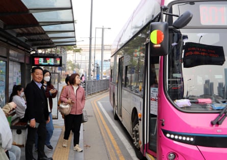 Kim Hooin waits at a bus stop with other communters next to a pink bus in Sejong, South Korea.