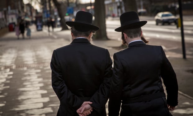 Two men in black hats, black coats and peyot walk along a pavement in Stamford Hill, London