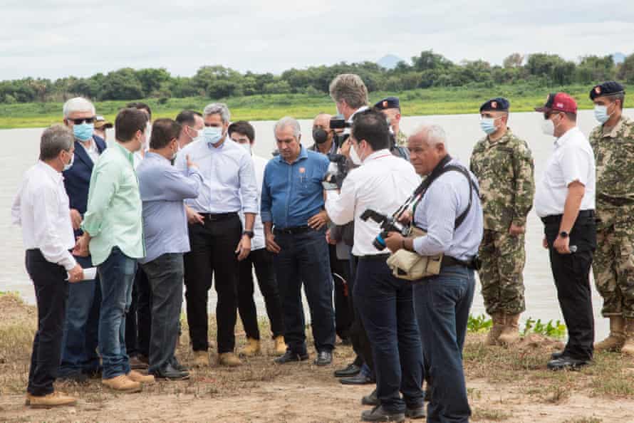 18 men, including photographers and army officers, stand by a wide river