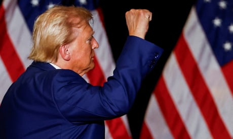 Donald Trump, wearing a blue suit, raises his fist at a rally, with American flags behind him