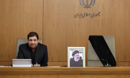 Iranian interim president dressed in black sits behind a desk next to a framed picture of the former president 