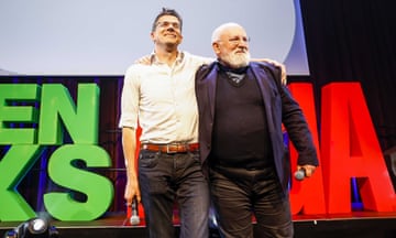 Bas Eickhout and Frans Timmermans hold microphones with their arms around each other's shoulders on a stage