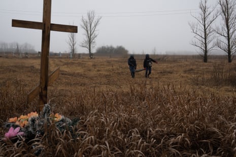 Two officers carry a shell across a field, with a wooden cross and flowers in the foreground