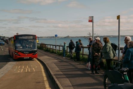 A queue of passengers stand next to the new bus stop by the sea, as a single-deck bus approaches