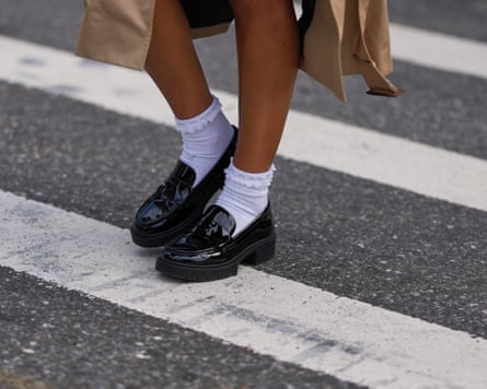 A closeup of someone’s calves and footwear. They wear a beige trenchcoat, black patent varnished leather chunky lug-sole loafers shoes worn with white crew socks, crossing a street with painted white markings.