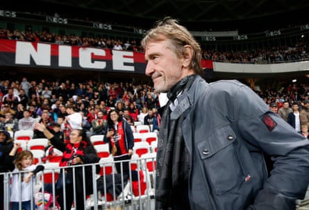 Ratcliffe wears a waxed jacket with a red and black Belstaff logo on the sleeve; he is on the pitch in Nice in front of football supporters.