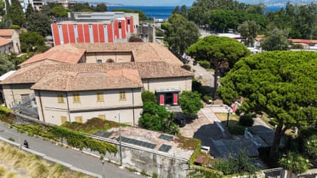A drone view of a brown roofed building between road and trees.