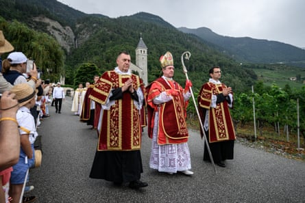 Procession of clerics in red robes with bishop at centre walking down crowd-lined road in mountainous region