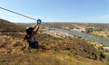 A woman with a ponytail and a helmet holds onto a zipwire while going downhill towards a river