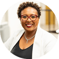 Smiling woman with short twists, red glasses, diamond necklace, and white blazer over black blouse
