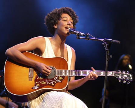A black woman in a long white dress plays the guitar and sings on stage.