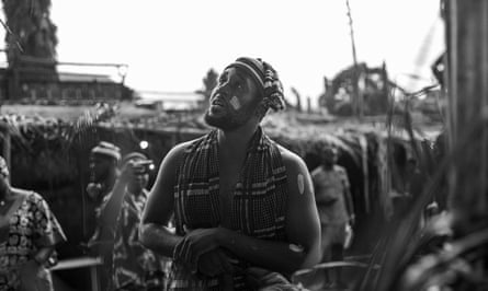 Black-and-white photo of a man in a scarf and hat with no shirt on, and paint on his face, gazing upwards