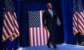 a man in a suit walks on stage in front of American flags