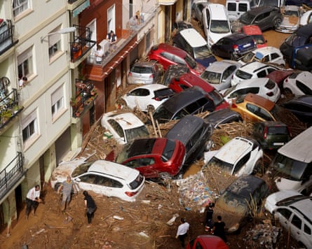 About 20 vehicles piled up as result of floods with several people by building in bottom left of image