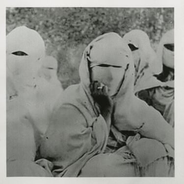 Several people in full-face white masks and hooded cloaks stand close together outdoors