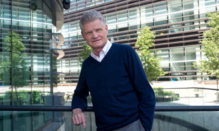 Sir John Bell leaning on a railing with a background of modern campus buildings with trees