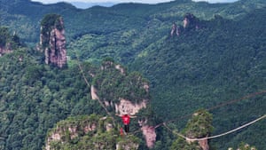 A man in a red T-shirt makes his way along a slackline high above a forested gorge with rocky outcrops