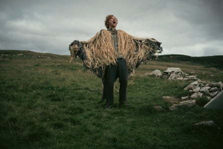 A man wearing a huge fringed shawl standing on a bleak moor witha dark sky behind him.
