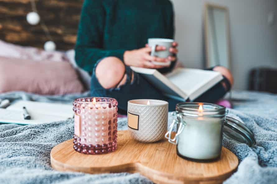 f a young woman relaxing with a book and a cup of coffee on her bed, behind three lit scented candles