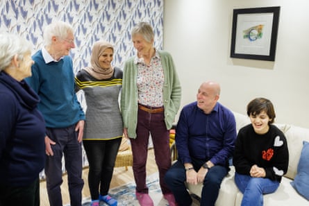 Two women and a man, all with grey hair, stand in a living room with a younger woman who wears a headscarf; another man sits on a sofa next to a teenage girl with collar-length dark hair, who wears jeans and a black baggy jumper with heart patterns. They are all smiling