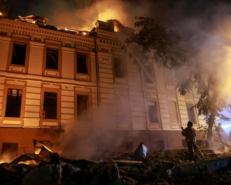 Firefighters work near a burning multi-story building at night, with smoke and debris visible