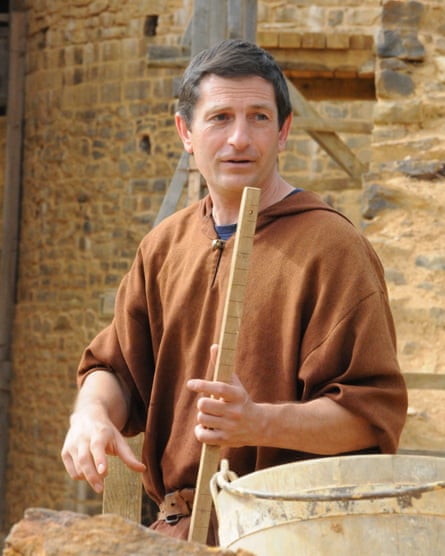 A man with short grey hair, wearing a brown hooded smock, wields a large stick on the building site of Chateau Guedelon