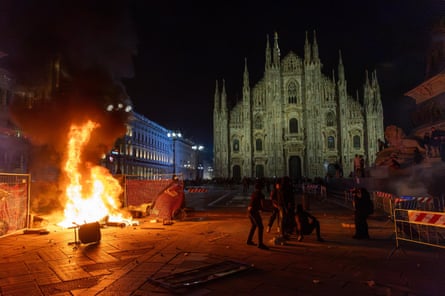 A pile of debris burns in the dark square with the cathedral illuminated in the background.
