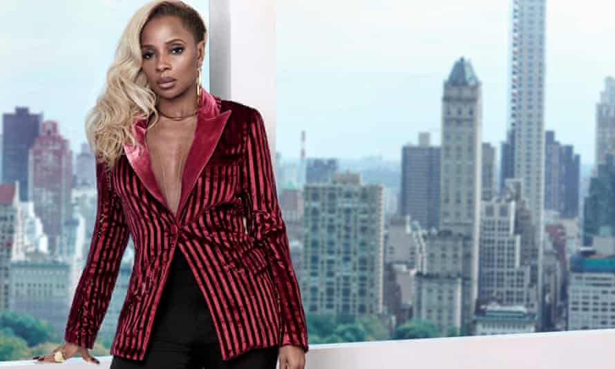 Mary J Blige, in a pink and black stripped jacket with a shiny pink lapel, standing in front of a window, the New York skyline behind her.