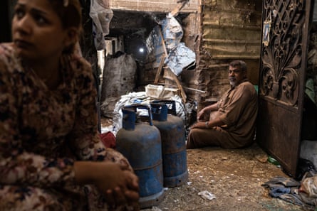 A man and woman sit in an enclosed space with a dirt floor. Two large gas canisters and bags of rubbish are next to them