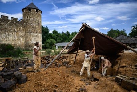 Outside in the grounds of Chateau Guedelon, two men in baggy 13th-century garb wield heavy hammers above rocks, shaded by tarpaulin, while another man rests his arm on a long stick and watches on