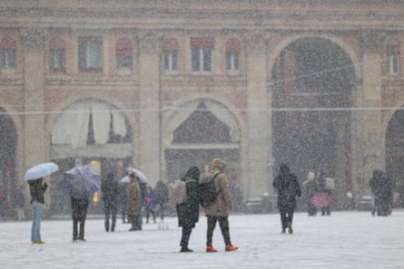 Snow falls as people stand or walk in a square in the city