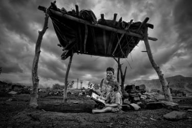 A young child sitting in the dirt reading a book, accompanied by an adult