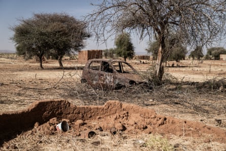 A burned out car and the remains of mud and brick houses in a dry-looking landscape with few trees.