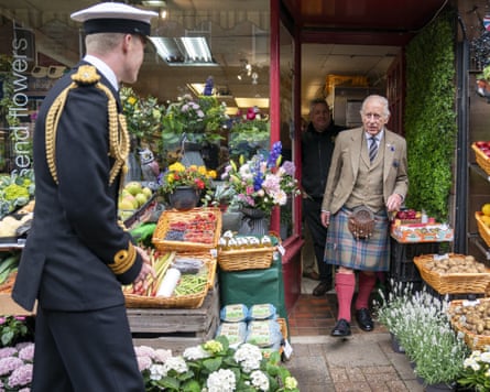 King Charles is walking out of a greengrocers. He is wearing a kilt, with a small amount of skin showing above his knee-high socks and below the bottom of the kilt