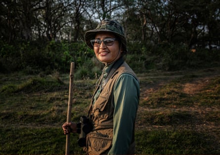 A woman in khaki clothing and a camouflage hat with a long stick smiles at the camera in a green outdoors setting