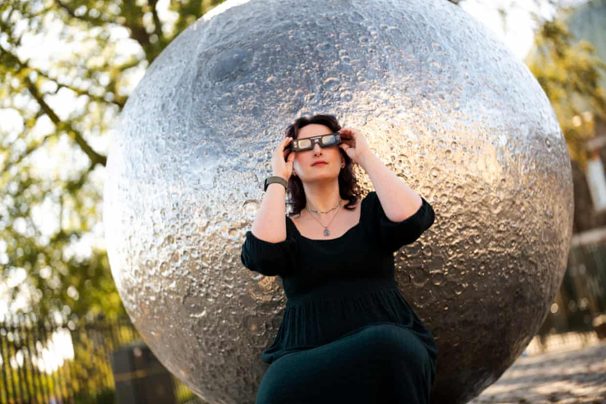 Dr Carys Hebert wears solar eclipse glasses while sitting in front a spherical silver-coloured sculpture