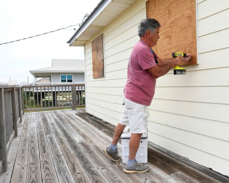 A man uses a drill to attach a sheet of plywood over the window of a wooden-board house