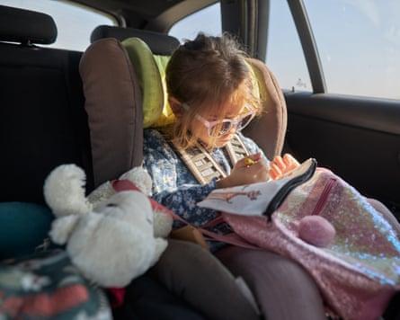 Young child wearing sunglasses and drawing in car seat on road trip.