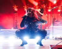 CMAT crouches low on stage holding a microphone, surrounded by red stage lighting and bandmates