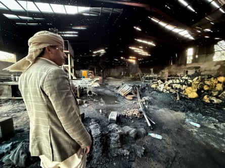 A man with a cream-coloured scarf wrapped around his head and a loose jacket surveys the scene inside an industrial building which appears to have been burned out, with debris piled to one wall.