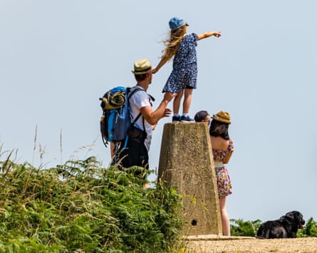 A young girl stands on a stone trig point looking out at the view with her family.