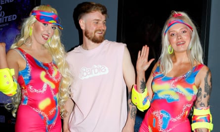 Two women dressed in blonde wigs and bright pink clothes on either side of bearded man wearing a Barbie T-shirt.